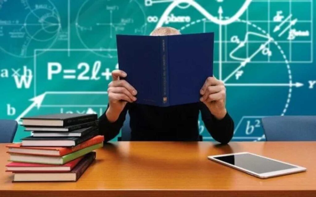 Un docente a scuola con volto coperto dal libro e dietro lavagna con numeri